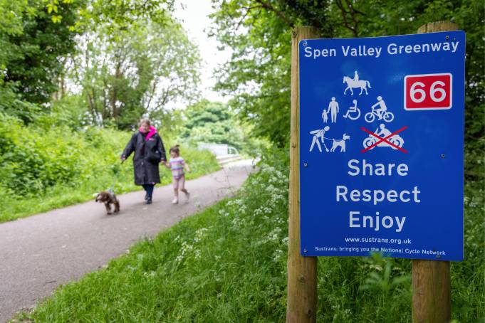 A woman and her daughter walk their dog along the Spen Valley Greenway. In the foreground, a sign says Share, Respect, Enjoy along with icons of what can and can't be done on the path