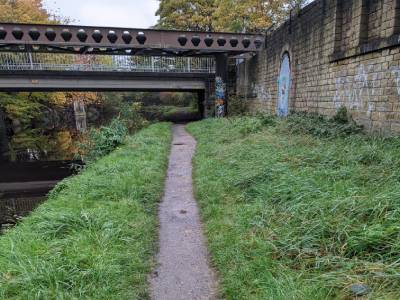 Slim gravel path running alongside the Huddersfield Broad Canal, bordered by water on one side and greenery on the other, with a bridge visible in the distance