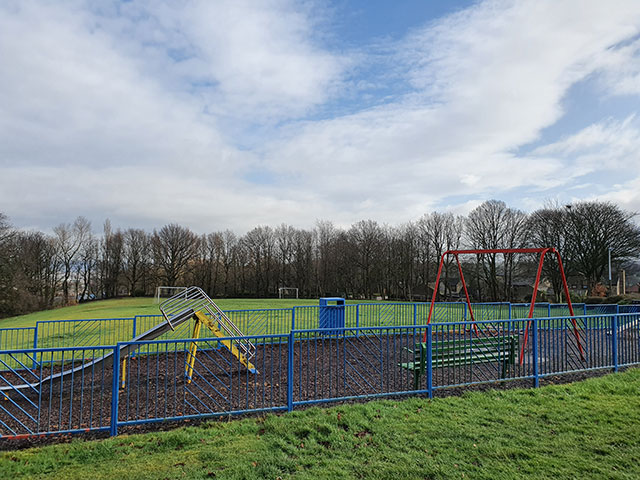 Upper Hopton Recreation Ground before regeneration. There is a swing and a slide and a bench. The play area is surrounded with a blue fence.